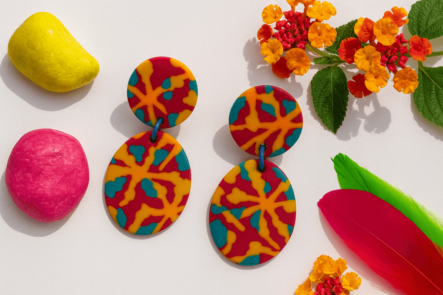 Red, teal and yellow polymer clay earrings with polymer clay pebbles, lantana flowers and bright feathers on a cream surface